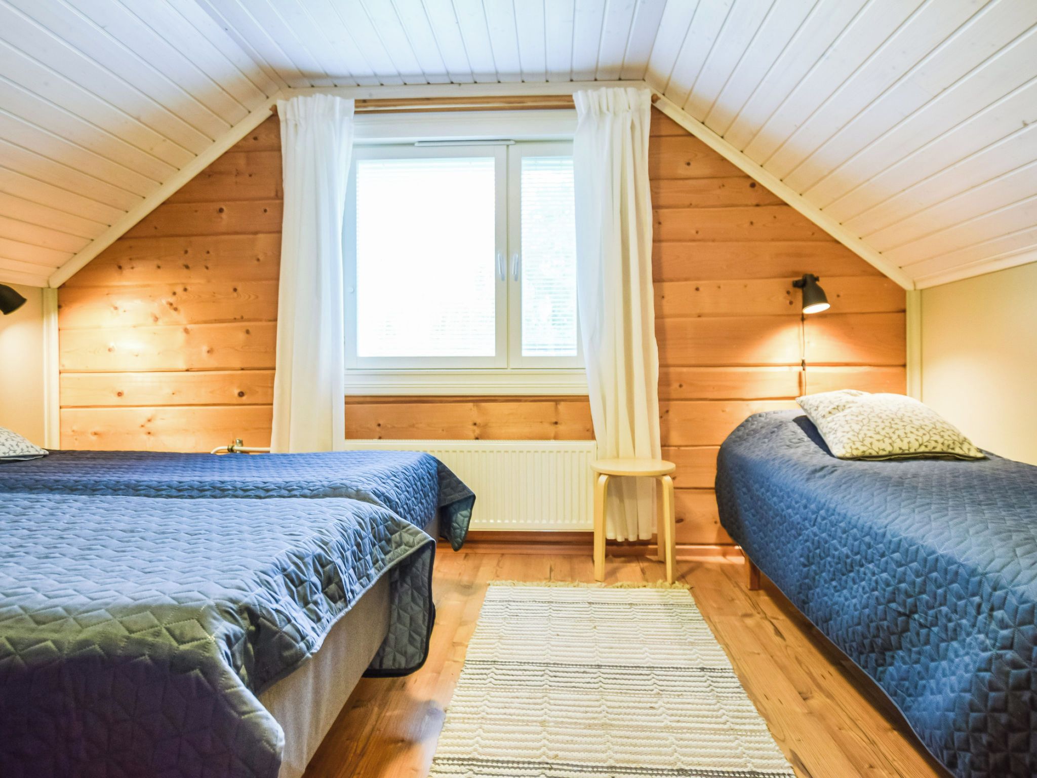 A cozy attic bedroom with two beds, a window with curtains, a small table, and a lamp.