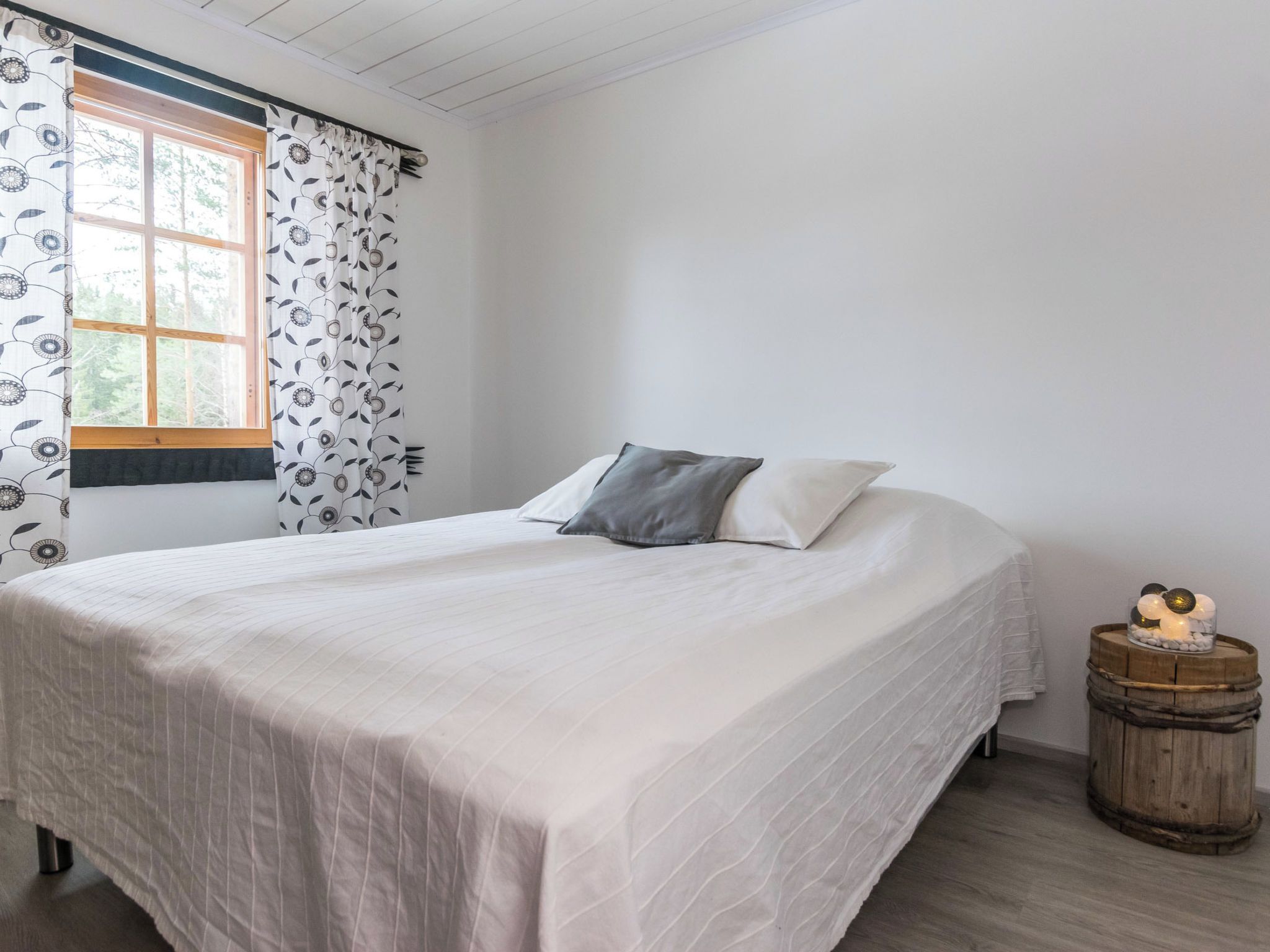 Ein Einzelbett mit weißer Bettdecke und zwei Kissen, ein Fenster mit gemusterten Vorhängen und ein kleiner Holzschemel mit dekorativen Gegenständen neben dem Bett.