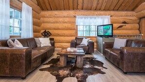 Interior de una cabaña de troncos con sofás de cuero, una mesa de centro de madera y un televisor.