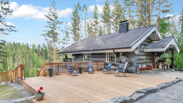 Una cabaña de troncos con terraza, mobiliario exterior y un bosque al fondo.