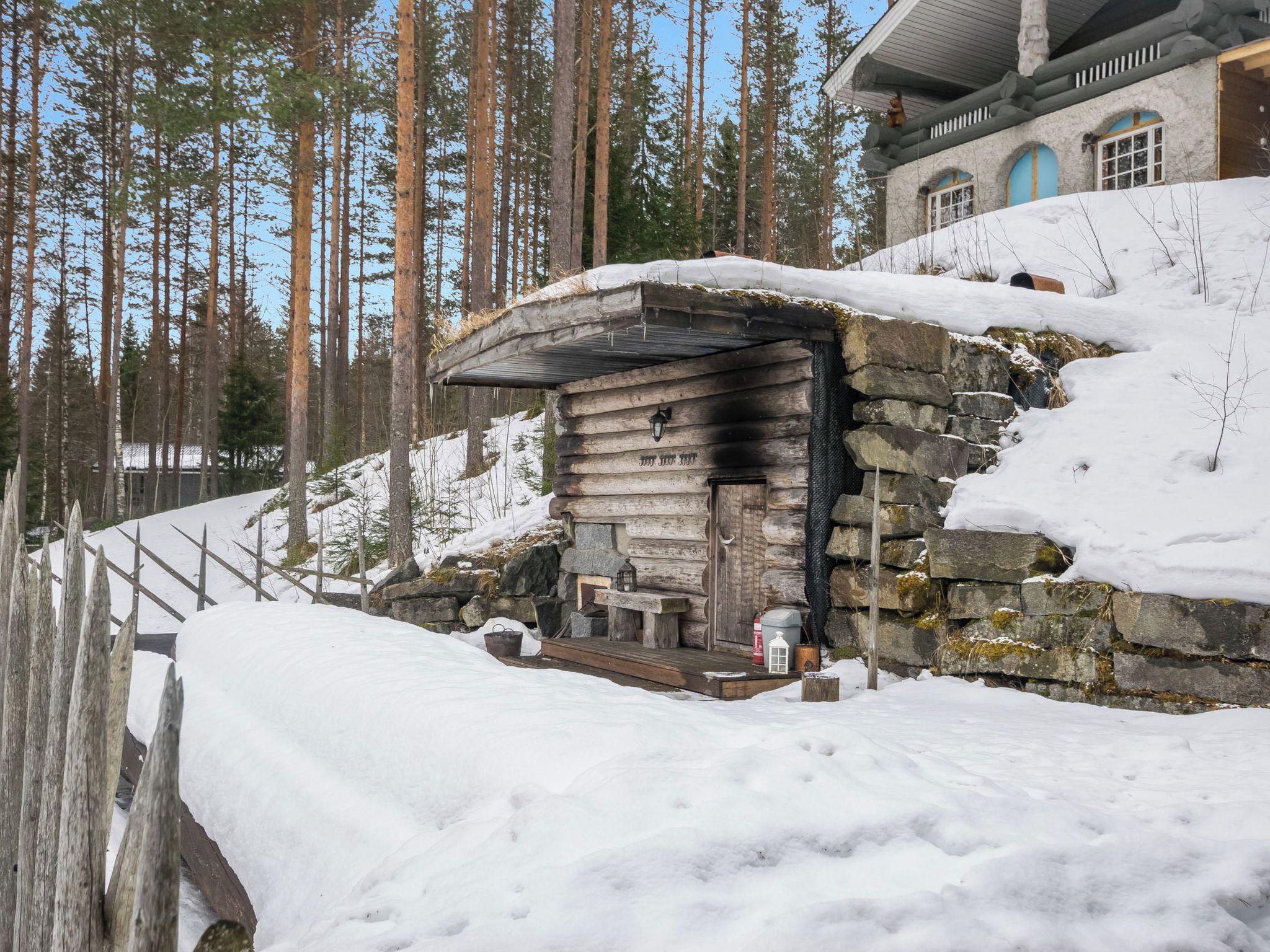 Hirsimökki, jossa on kivimuuri, penkki ja lyhty ulkona.