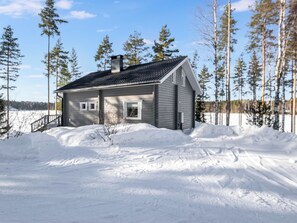 Una casa de un piso con techo oscuro y revestimiento claro, rodeada de nieve y árboles.