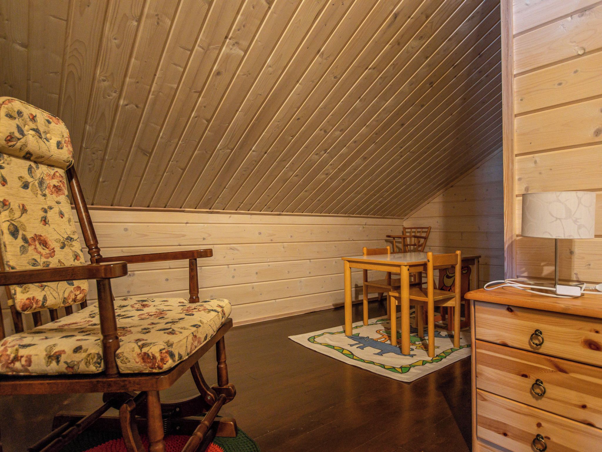 Een gezellige kamer onder de schuine kap met houten wanden. Er staat een schommelstoel met bloemenprint, een houten tafel met stoelen en een kledingkast met lades.
