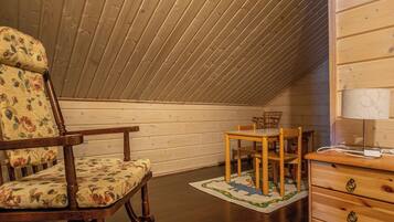 Een gezellige kamer onder de schuine kap met houten wanden. Er staat een schommelstoel met bloemenprint, een houten tafel met stoelen en een kledingkast met lades.