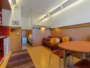 Un espacio habitable compacto con kitchenette, un sofá cama, una mesa de comedor con sillas y una alfombra estampada.