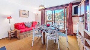 A cozy living room with a red sofa, a dining table with chairs, a floor lamp, and a window with curtains. - Vostok Zodiaque by Interhome (Le Corbier)
