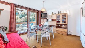 A cozy living room with a red sofa, a dining table with patterned chairs, a TV, and a cabinet with glass doors. - Vostok Zodiaque by Interhome (Le Corbier)