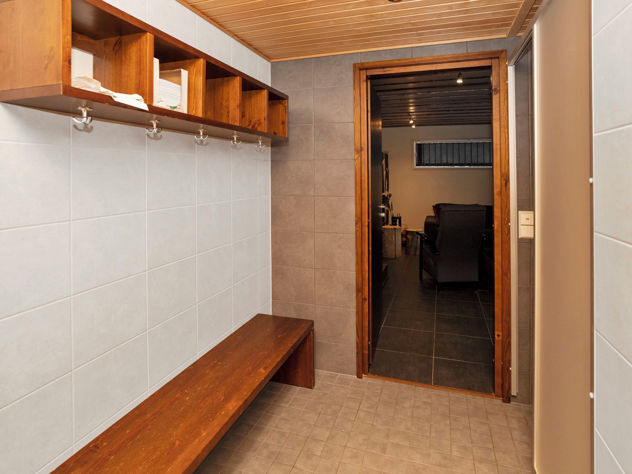 A bathroom with a wooden bench, tiled walls, and a doorway leading to a room with a chair and a table.