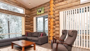 L’intérieur d’une cabane en rondins est aménagé avec un canapé d’angle, une table basse en bois et une grande fenêtre donnant sur des arbres couverts de neige.