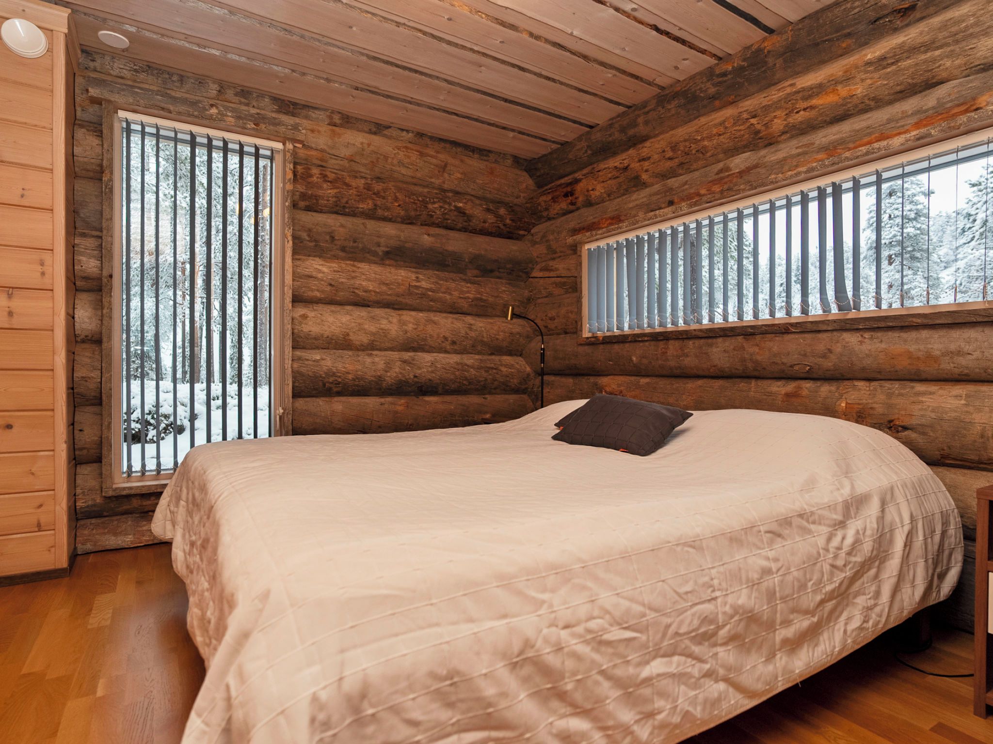 A log cabin bedroom with a bed, two windows, and a wooden ceiling.