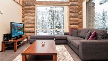 A log cabin interior with a sectional sofa, a wooden coffee table, a television on a wooden stand, and a large window with a snowy view.