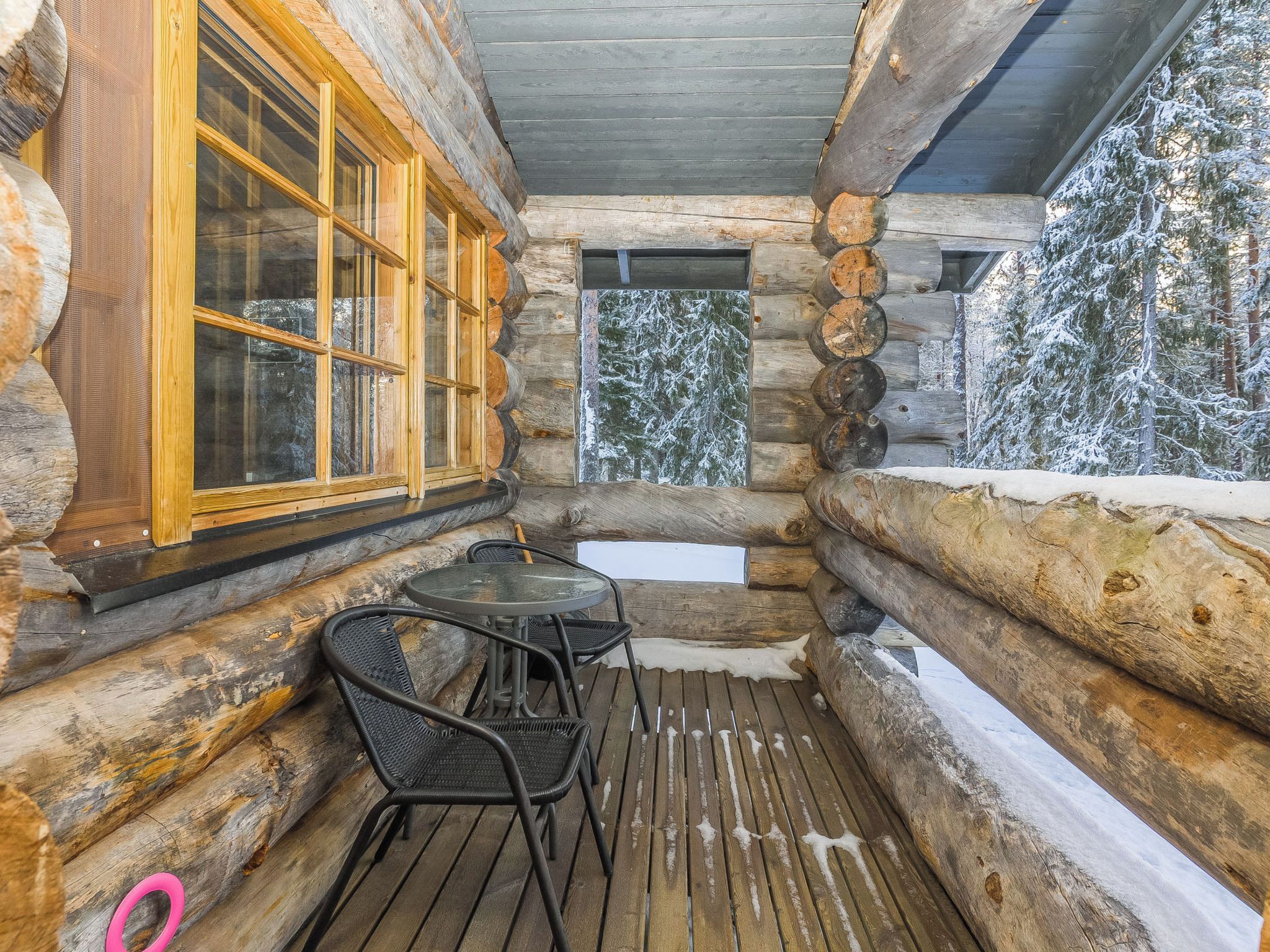 A log cabin with a wooden deck, a table with chairs, and a window with a view of snow-covered trees.
