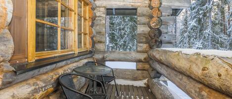 A log cabin with a wooden deck, a table with chairs, and a window with a view of snow-covered trees.
