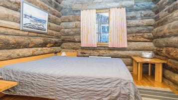 A log cabin bedroom with a bed, a small table, a window with curtains, and a framed picture on the wall.