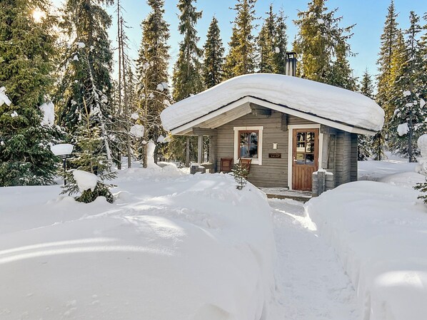 A small cabin with a snow-covered roof, surrounded by snow and trees.
