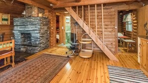 A wooden cabin interior with a stone fireplace, wooden floors, a staircase, and a dining area.