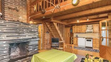 A rustic cabin interior with a stone fireplace, a wooden staircase, a kitchen area, and a dining table with a green tablecloth.