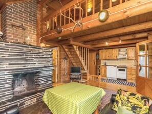 A rustic cabin interior with a stone fireplace, a wooden staircase, a kitchen area, and a dining table with a green tablecloth. - Tallukka by Interhome (Pertunmaa)