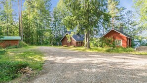 A gravel driveway leading to a cabin with a metal roof and a chimney, surrounded by tall trees and a small shed.