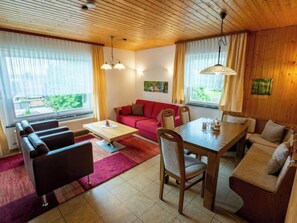 A cozy living room with a red sofa, a wooden dining table, and a coffee table. There are two windows with curtains, a radiator, and a ceiling with wooden beams. - Vacation home Romeo by Interhome (Dittishausen)