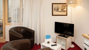 Een hotelkamer met een leren fauteuil, een televisie op een standaard, een tafeltje met een afstandsbediening, een lijst met een afbeelding aan de muur, en een raam met gordijnen.