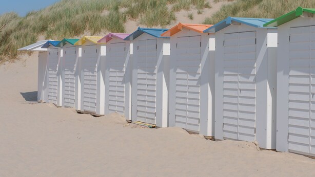 Una fila di casette sulla spiaggia con pareti bianche e tetti colorati, affacciata su una spiaggia sabbiosa con dune sullo sfondo.