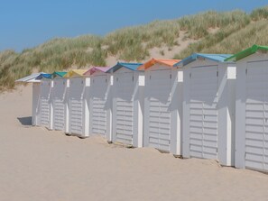 A row of beach huts with white walls and colorful roofs, situated on a sandy beach with dunes in the background.