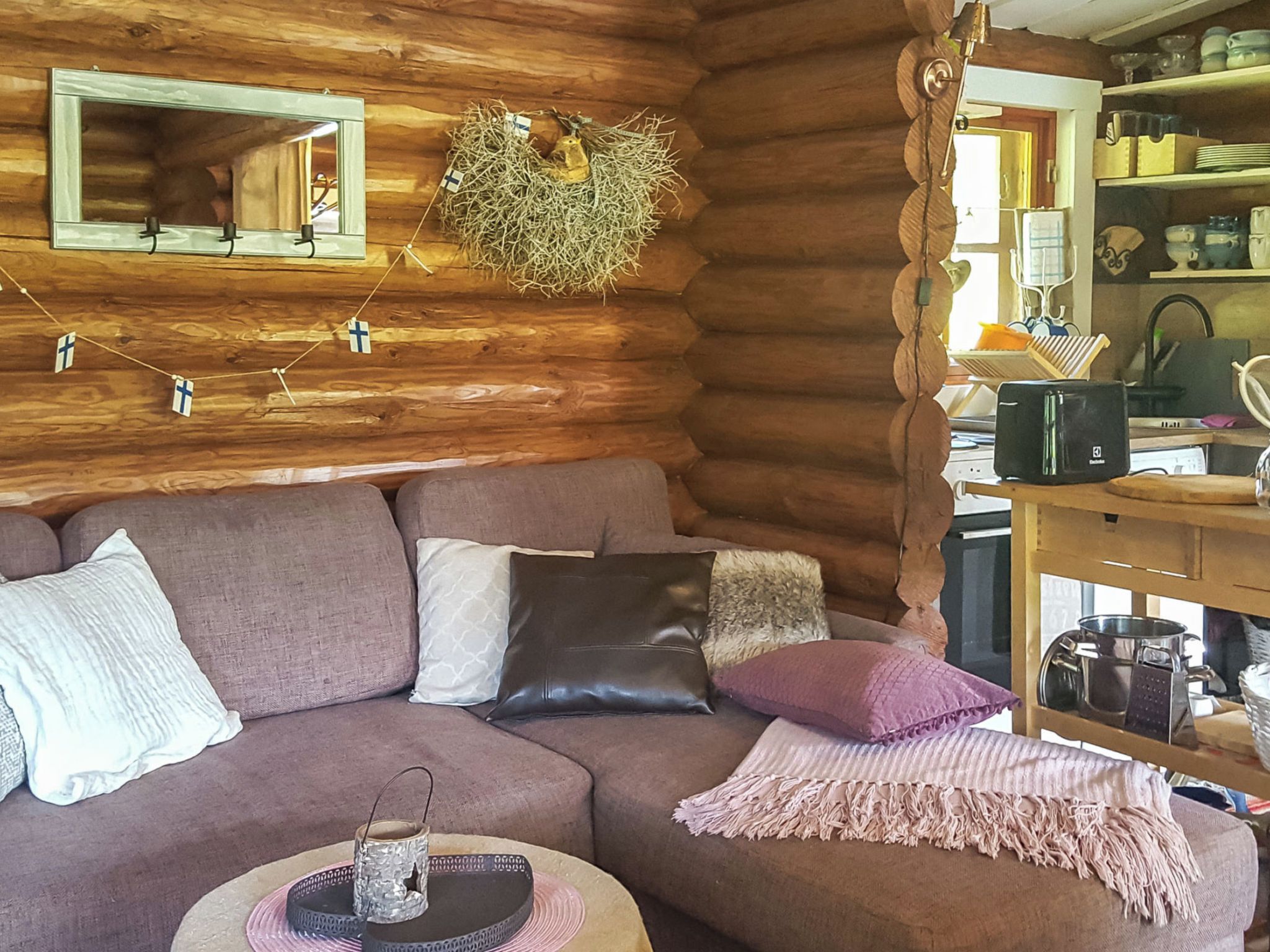 A log cabin interior with a wooden wall, a sofa with cushions, a small table with a candle, and a kitchen area with a stove and utensils.