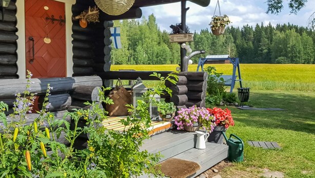 Una cabana de fusta amb un porxo de fusta, una porta vermella i una zona de jardí amb diverses plantes i flors.