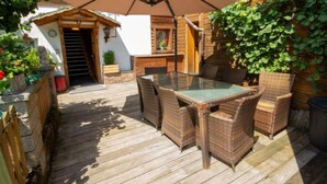 Een houten terras met een glazen tafel en rieten stoelen, omgeven door een houten hek en groen.