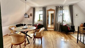 A cozy, well-lit room with a wooden floor, a round dining table, wicker chairs, a hanging light fixture, and a view of a green garden through a window with curtains. - Hiddensee by Interhome (Dargun)