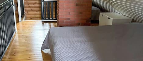 Een slaapkamer met een schuine kap, houten vloer, een bed, een raam en een open haard van baksteen.