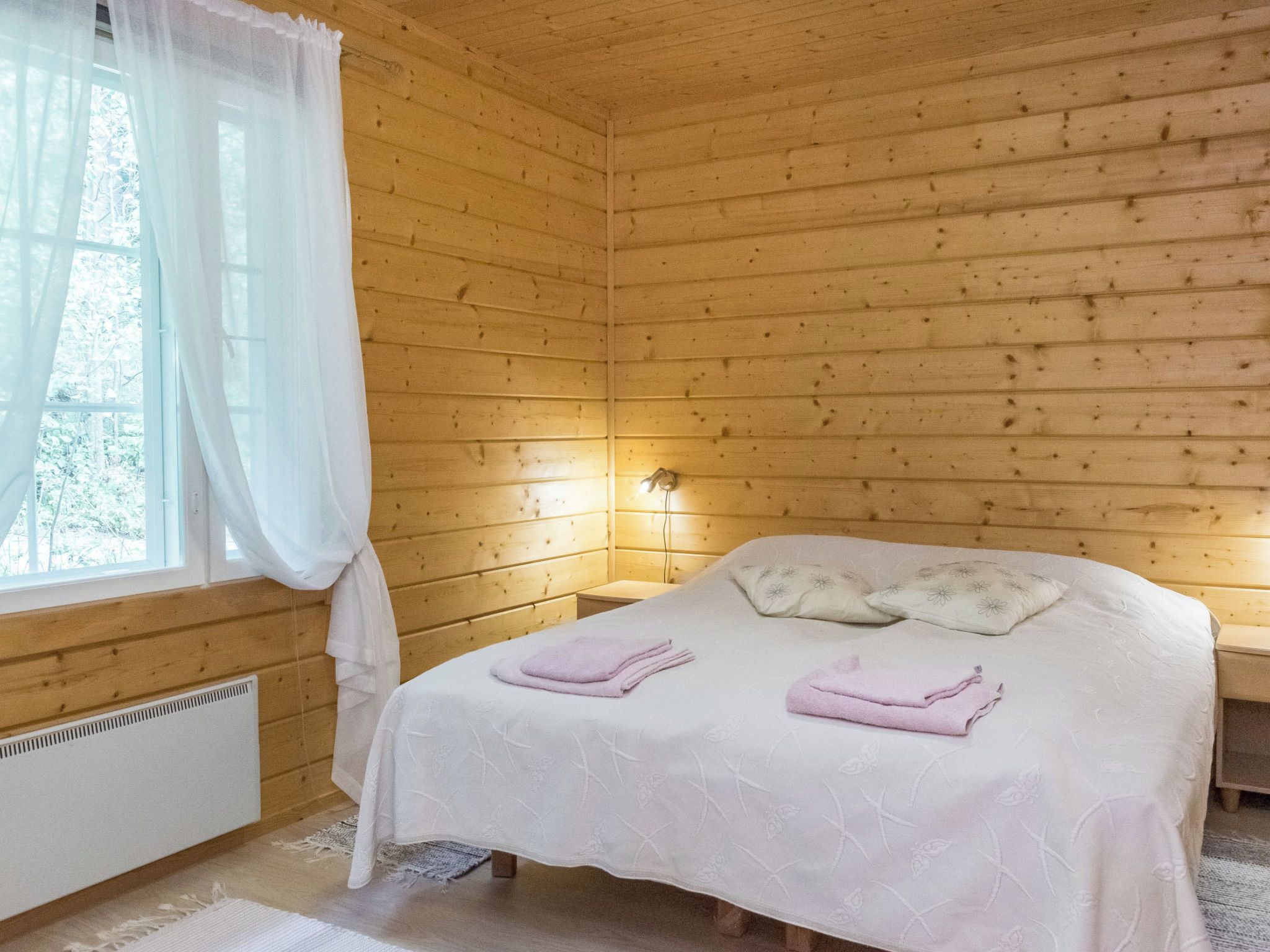 A wooden cabin room with a bed, a nightstand, a window with curtains, and a wall-mounted light.