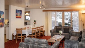 Una sala acogedora con sofás a cuadros, un comedor con una mesa dispuesta para dos personas y vistas a árboles nevados a través de amplias ventanas.