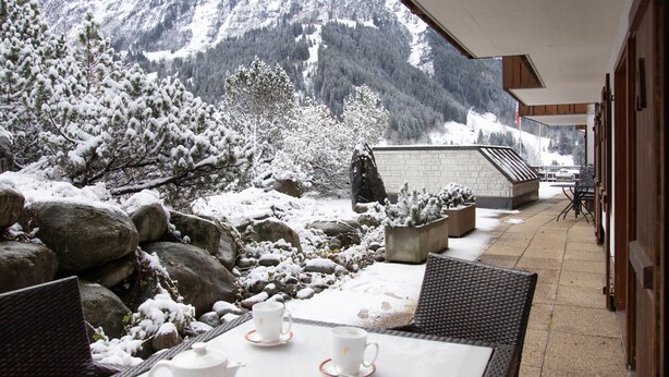 Un área de estar al aire libre cubierta de nieve con vista a la montaña, que incluye una mesa con tetera y tazas, y plantas en macetas.