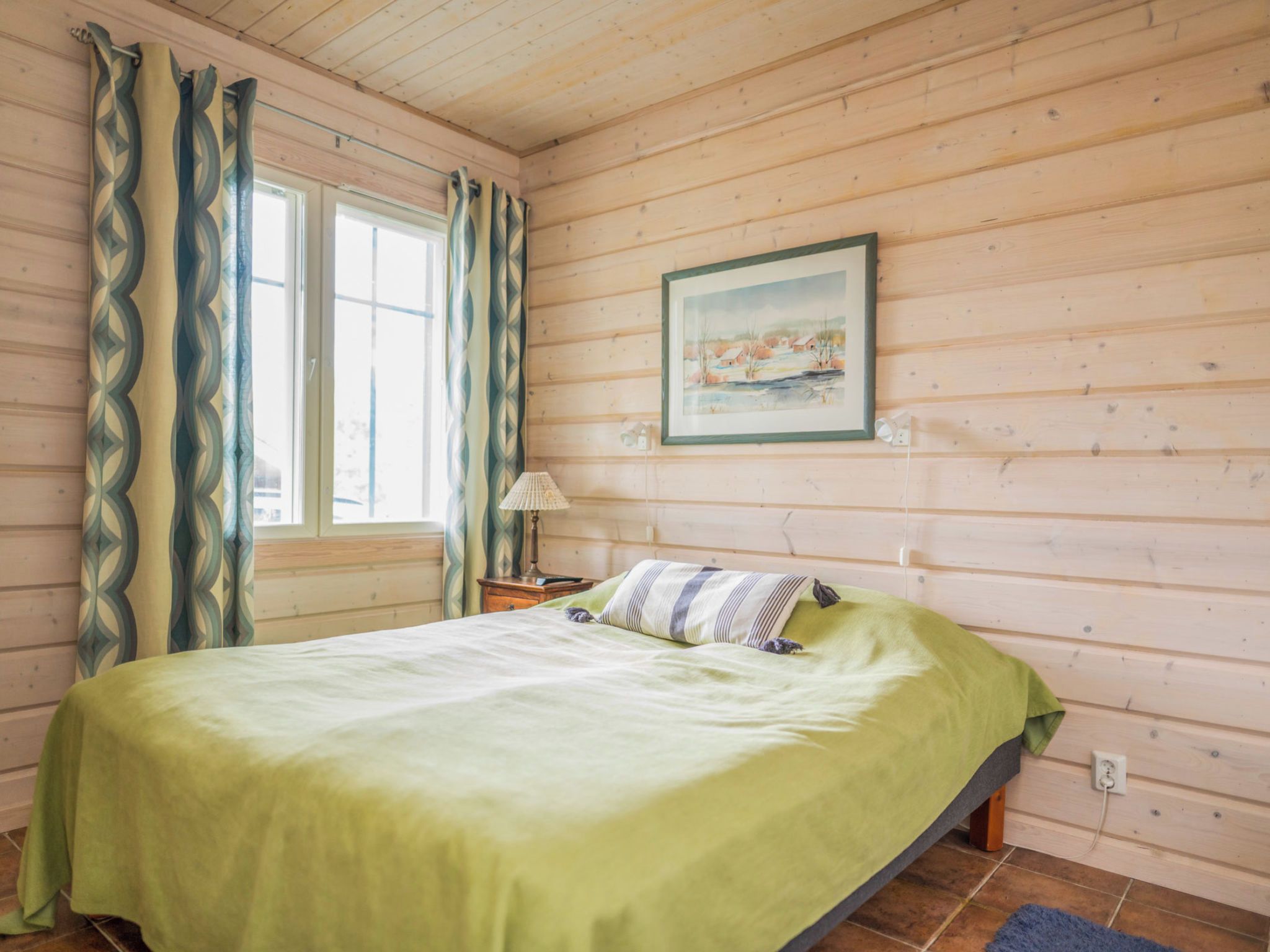 Una habitación con cama, ventana con cortinas, un cuadro en la pared y lámpara en la mesita de noche.