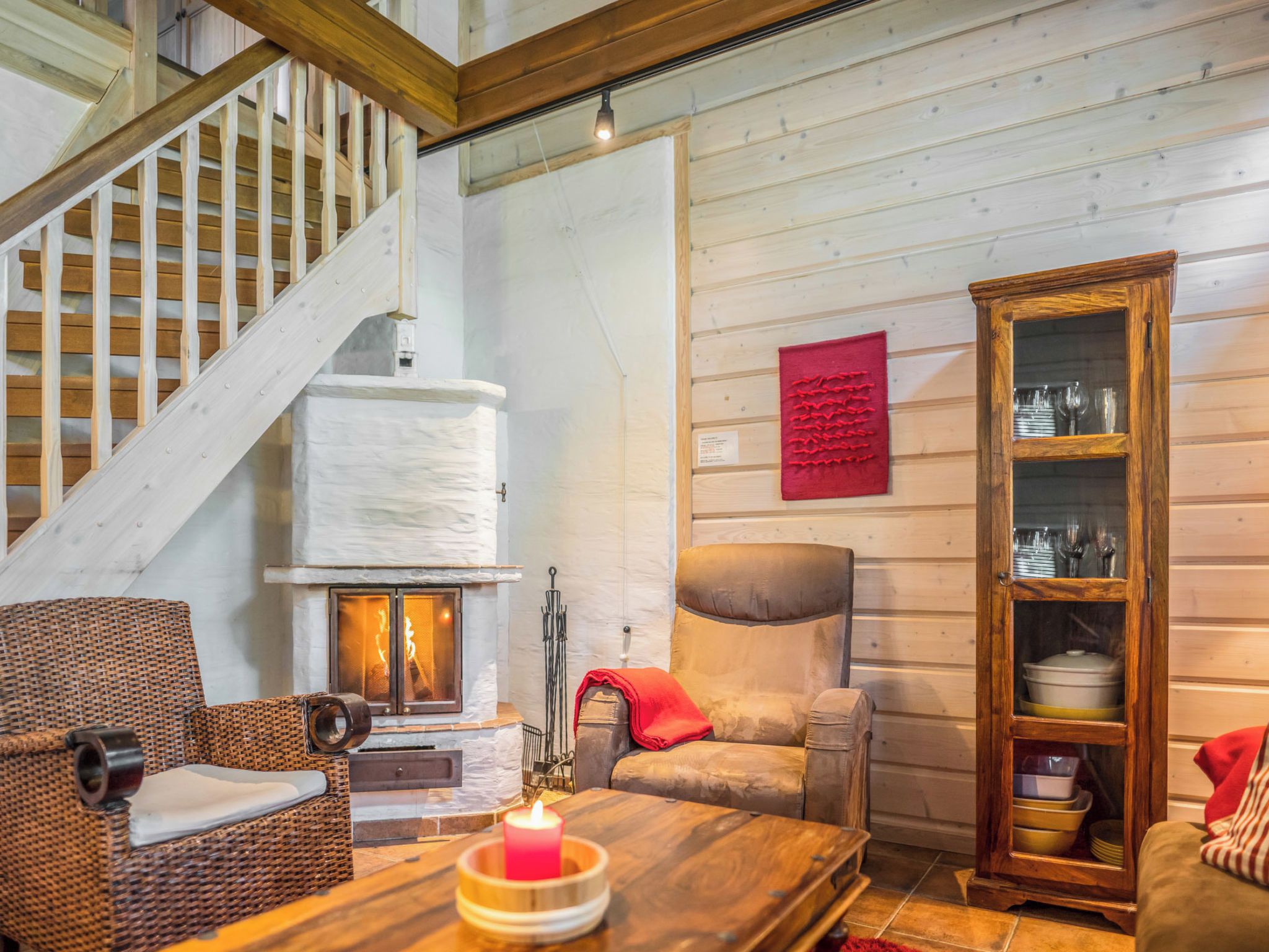 Un interior acogedor con chimenea, una escalera de madera, un sillón de mimbre, un sofá con cojines y un mueble de madera con puertas de cristal.