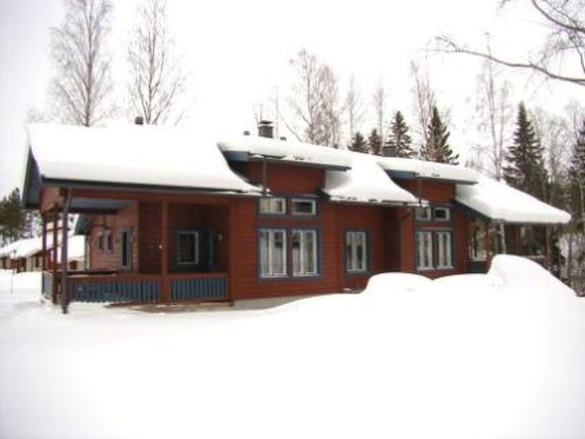 Una cabaña de madera con porche y techo cubierto de nieve, rodeada de árboles.