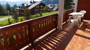 A balcony with a wooden railing, a table with two chairs, and a view of a mountain landscape.