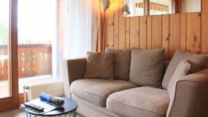 A cozy room with a wooden wall, a beige sofa, a small round table with a remote control, and a window with curtains.