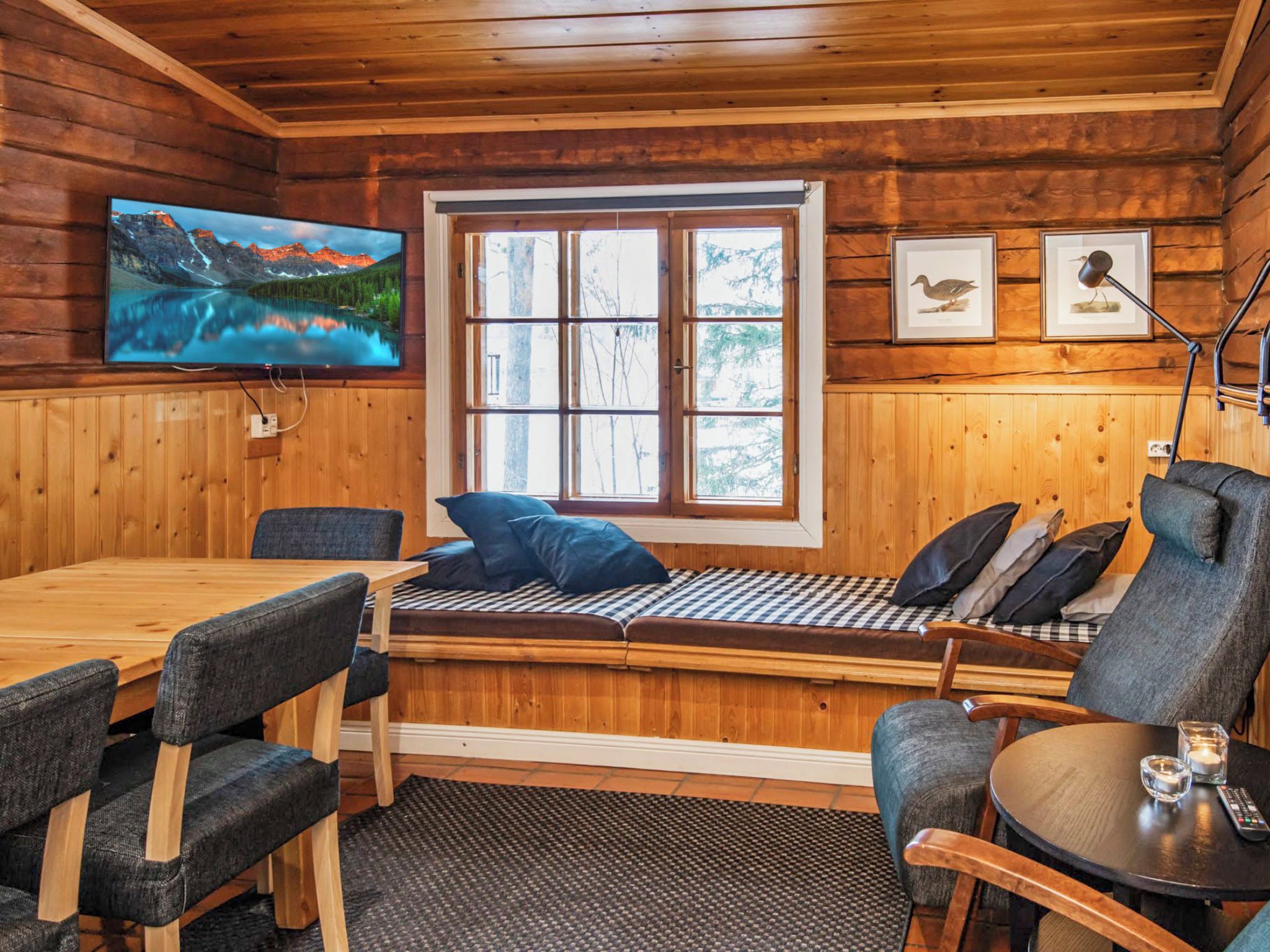 A cabin interior with a wooden dining table, chairs, a bench with cushions, a television mounted on the wall, and framed pictures.