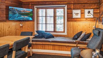 A cabin interior with a wooden dining table, chairs, a bench with cushions, a television mounted on the wall, and framed pictures.