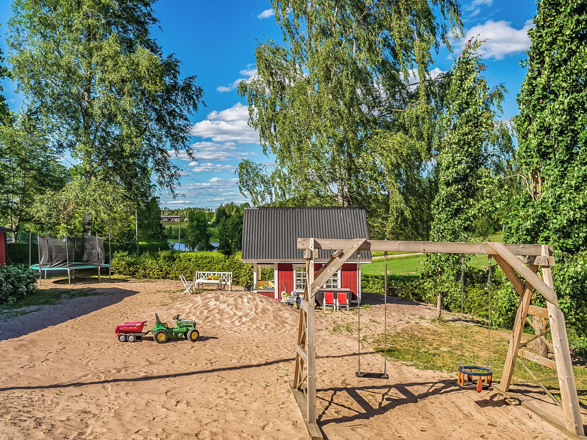 Un espace extérieur sablonneux avec une balançoire, un petit bâtiment rouge et un tracteur vert.