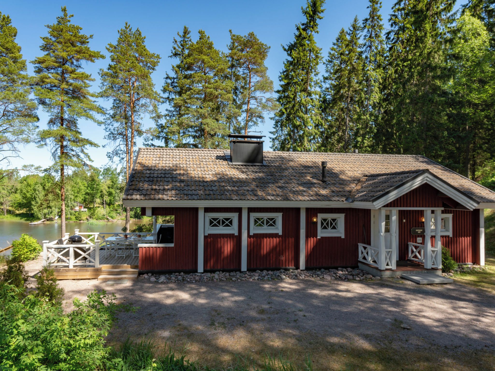 Une cabane rouge avec un toit en tuiles, une terrasse et une vue sur un lac entouré d’arbres.