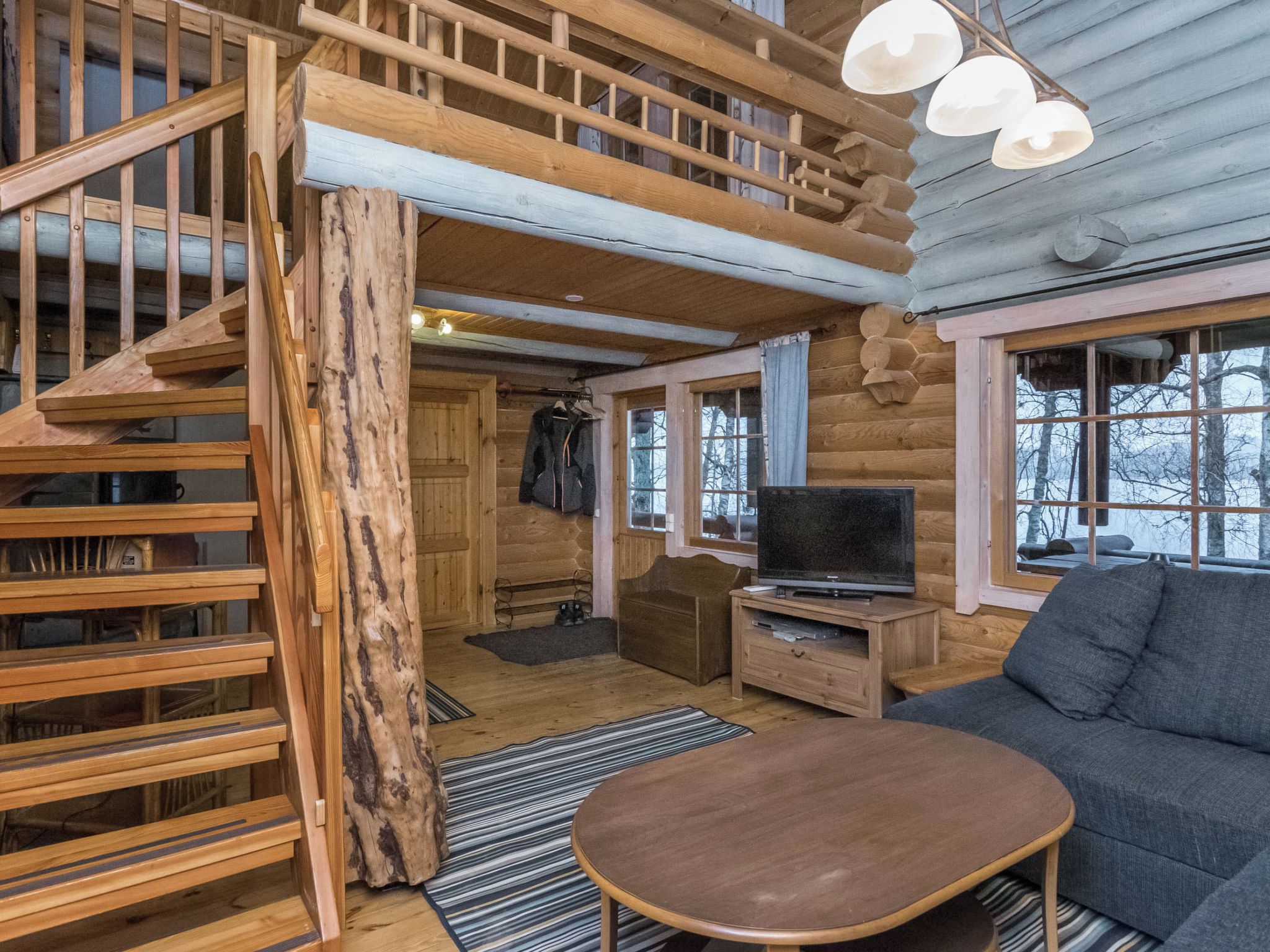 Interior de una cabaña de troncos con escalera de madera, televisor, sofá y un gran ventanal.