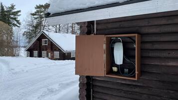 Une cabane en bois, dont le toit est recouvert de neige, et sur la paroi extérieure de laquelle est fixée une borne de recharge pour véhicules Tesla.