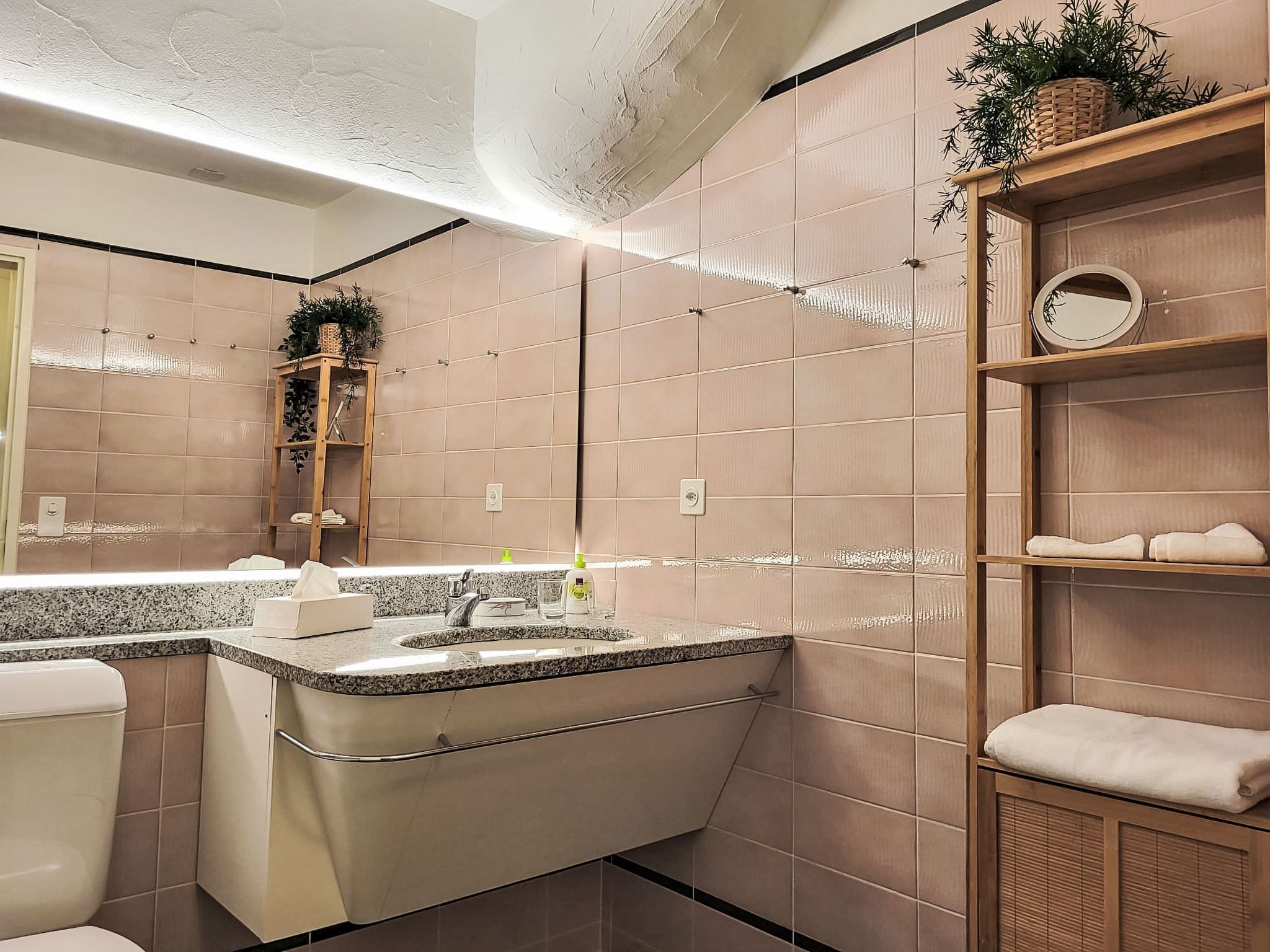 A modern bathroom with a vanity, sink, and toilet. There are shelves with towels and a potted plant.