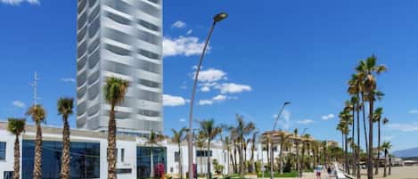 Ein modernes Hotel mit geschwungener Fassade, einem gepflegten Garten und einer Strandpromenade gesäumt von Palmen.