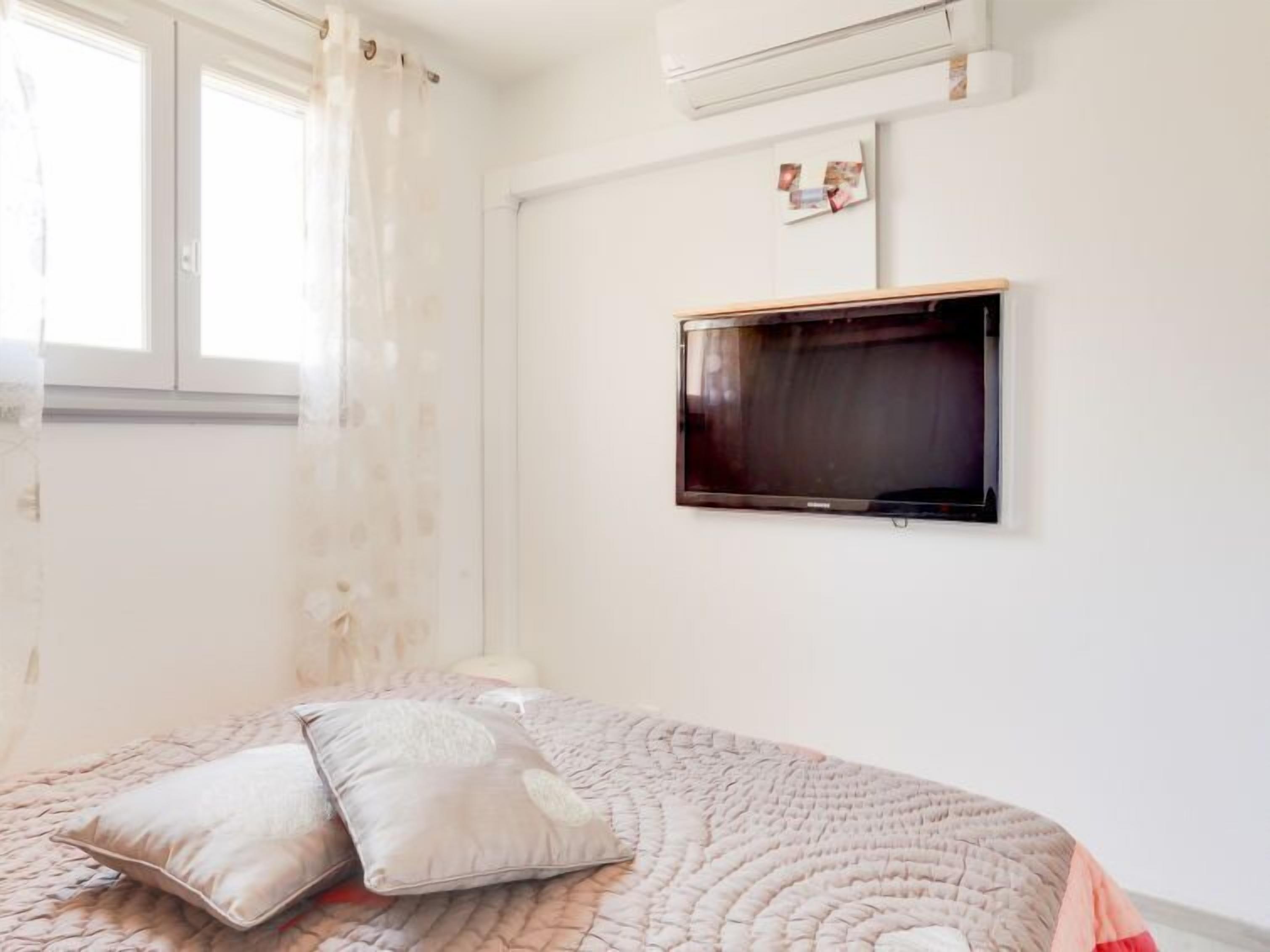 Een slaapkamer met een bed, kussens, een aan de muur bevestigde televisie en een raam met gordijnen.
