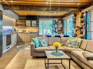 A modern kitchen with wooden accents, a dining area with a sofa and coffee table, and a view through large windows.